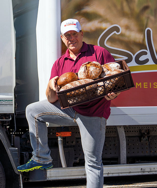 Das Bild zeigt einen Mitarbeiter von Scherf vor einem Lieferwagen, der einen großen, gefüllten Brotkorb in beiden Händen hält.